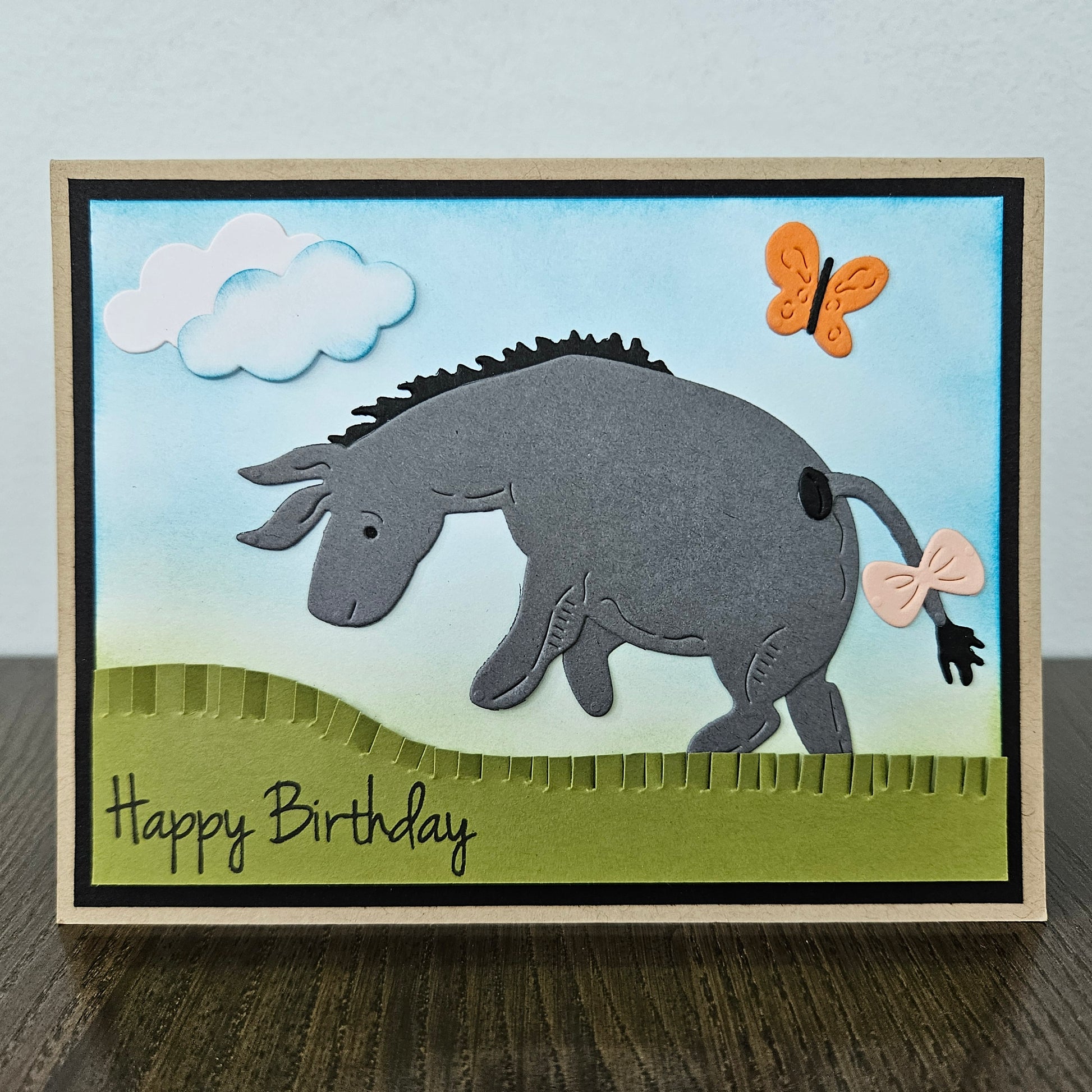 Front of a handmade birthday card showing a 3D layered paper-pieced donkey inspired by Eeyore with a pink bow on his tail, set against a blue inked sky with clouds, an orange butterfly, and a green grass hill.