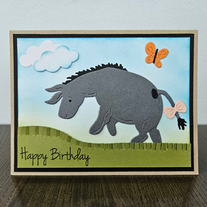 Front of a handmade birthday card showing a 3D layered paper-pieced donkey inspired by Eeyore with a pink bow on his tail, set against a blue inked sky with clouds, an orange butterfly, and a green grass hill.