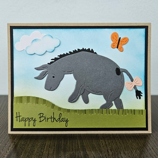 Front of a handmade birthday card showing a 3D layered paper-pieced donkey inspired by Eeyore with a pink bow on his tail, set against a blue inked sky with clouds, an orange butterfly, and a green grass hill.