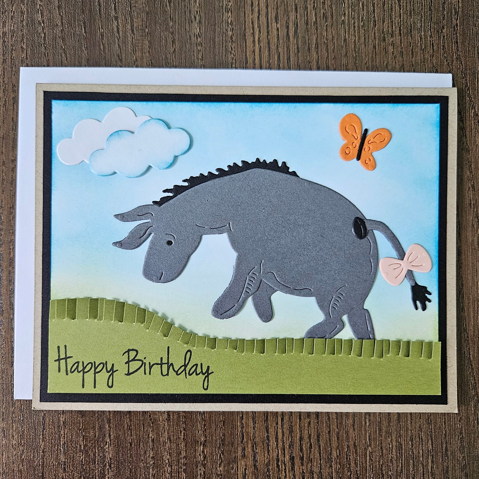 Front of a handmade birthday card showing a 3D layered paper-pieced donkey inspired by Eeyore with a pink bow on his tail, set against a blue inked sky with clouds, an orange butterfly, and a green grass hill.