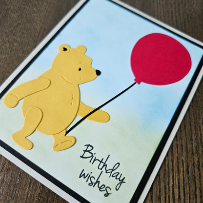 Card with a bear holding a red balloon and 'Birthday wishes' text on a wooden surface