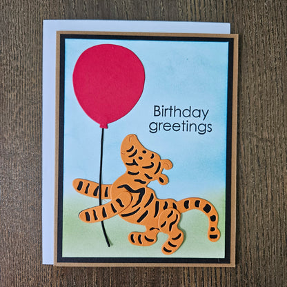 Handmade Winnie the Pooh birthday card featuring a die-cut illustration of Tigger holding a bright red balloon against a soft hand-inked watercolor sky and green meadow background.