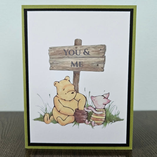 Handmade Winnie the Pooh friendship card featuring a classic illustration of Pooh and Piglet sitting together beneath a rustic wooden sign that reads You and Me, framed by a distinct black inner frame and a sage green layered border.
