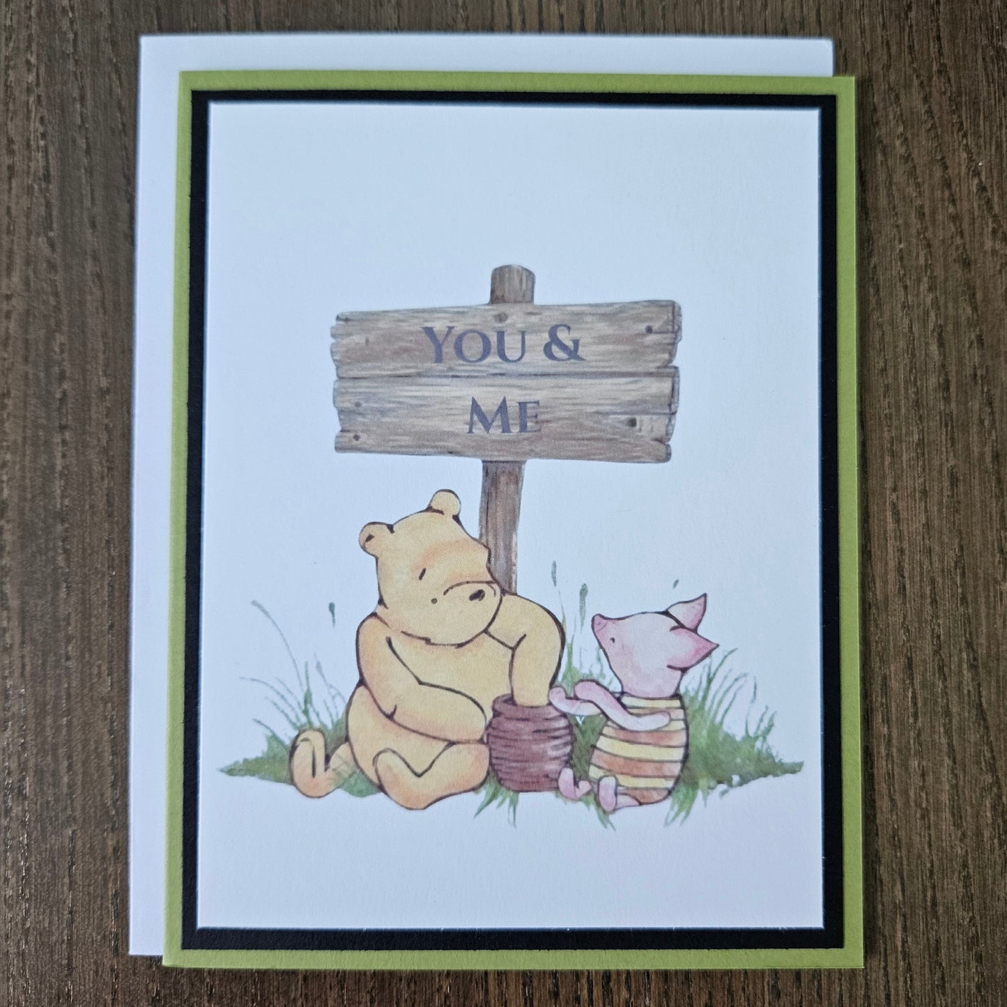Handmade Winnie the Pooh friendship card featuring a classic illustration of Pooh and Piglet sitting together beneath a rustic wooden sign that reads You and Me, framed by a distinct black inner frame and a sage green layered border.