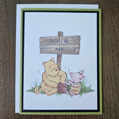 Handmade Winnie the Pooh friendship card featuring a classic illustration of Pooh and Piglet sitting together beneath a rustic wooden sign that reads You and Me, framed by a distinct black inner frame and a sage green layered border.