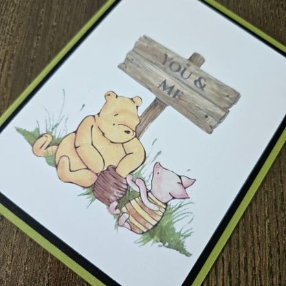 Handmade Winnie the Pooh friendship card featuring a classic illustration of Pooh and Piglet sitting together beneath a rustic wooden sign that reads You and Me, framed by a distinct black inner frame and a sage green layered border.