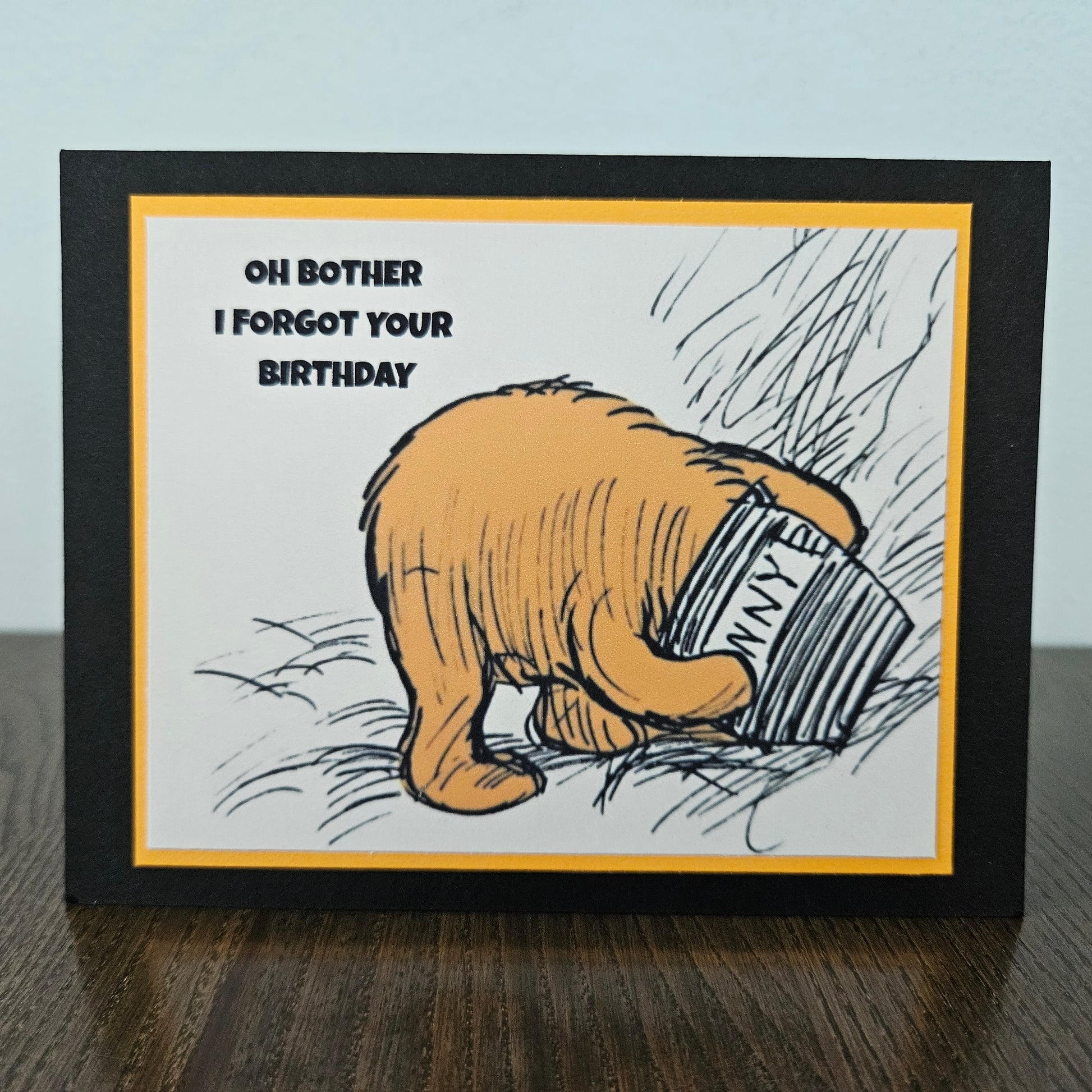 Card with cartoon bear holding a book titled 'Oh Bother I Forgot Your Birthday' on a wooden surface.