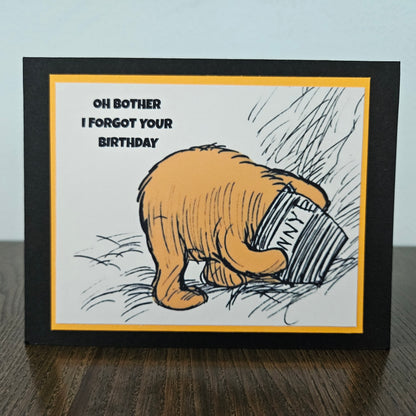 Card with cartoon bear holding a book titled 'Oh Bother I Forgot Your Birthday' on a wooden surface.