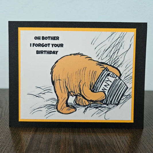 Card with cartoon bear holding a book titled 'Oh Bother I Forgot Your Birthday' on a wooden surface.