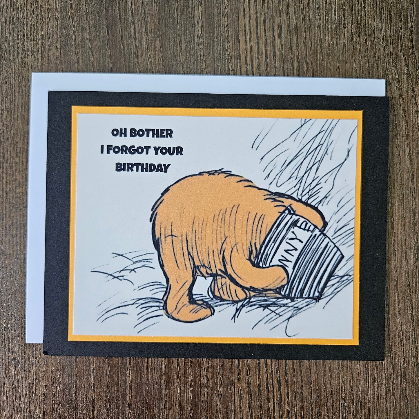 Card with a hand holding a can and text 'Oh bother I forgot your birthday' on a wooden surface