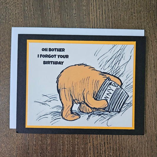 Card with a hand holding a can and text 'Oh bother I forgot your birthday' on a wooden surface