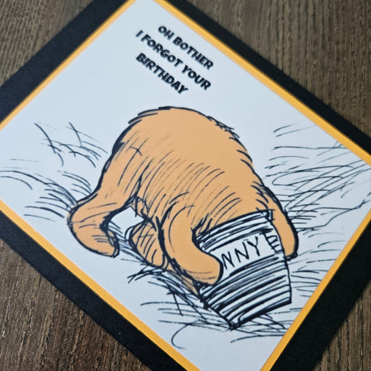 Card with a hand holding a box labeled 'Mummy' and text 'Oh bother I forgot your birthday' on a wooden surface.