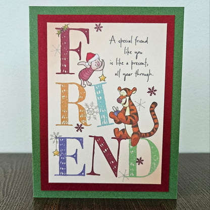 A handmade greeting card featuring a Winnie the Pooh themed design with the word 'FRIEND' in large decorative letters, a pig in a Santa hat, Tigger, and a handwritten message, set against a green and red cardstock border on a wooden surface.