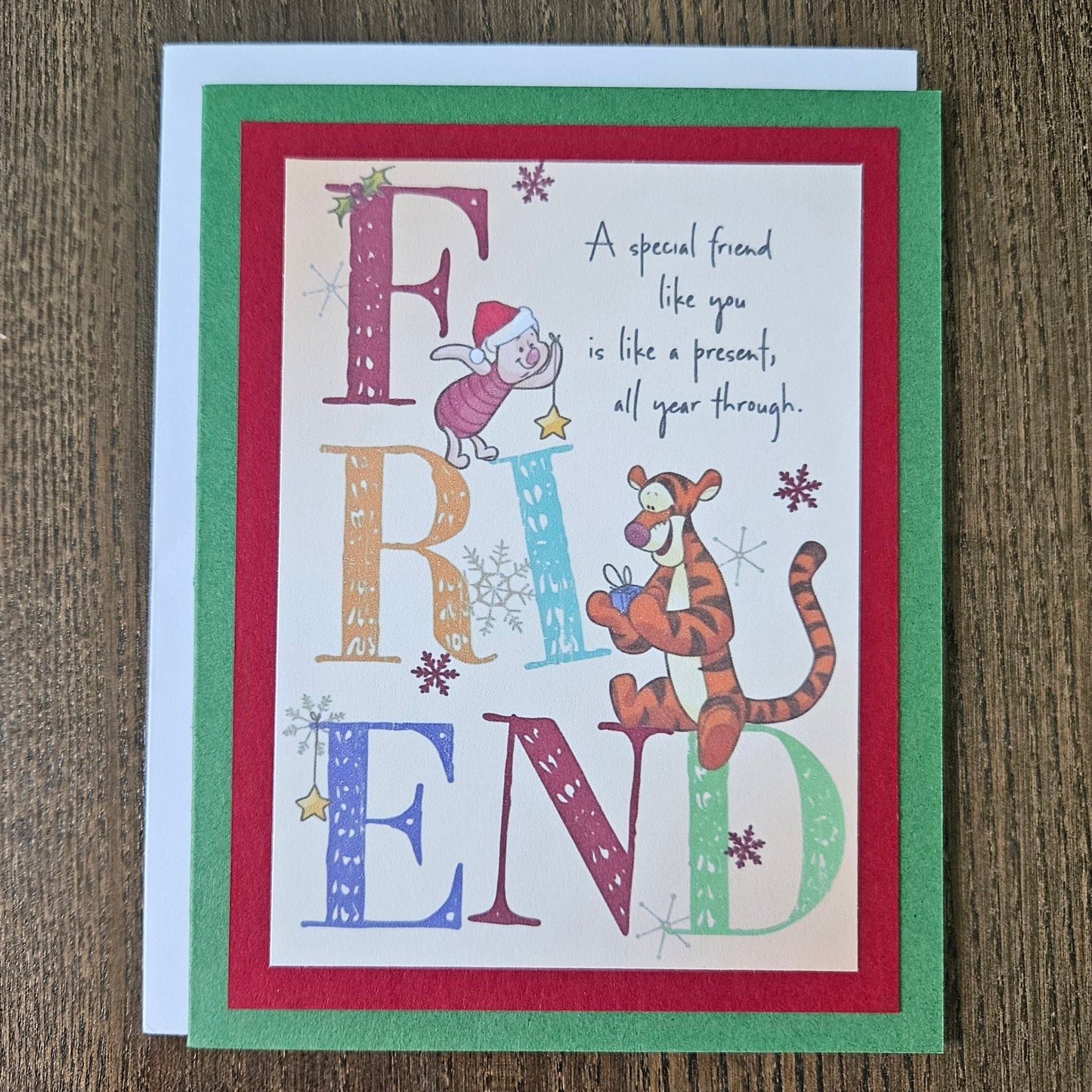 A square greeting card featuring Winnie the Pooh and Tigger with the word 'FRIEND' spelled out in large decorative letters, placed on a white envelope over a brown wood surface.