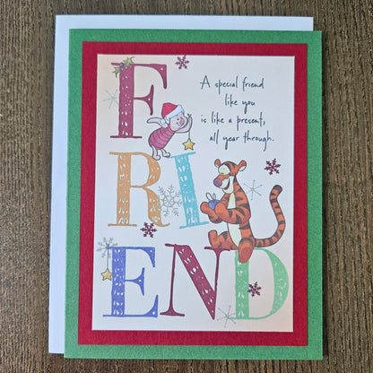 A square greeting card featuring Winnie the Pooh and Tigger with the word 'FRIEND' spelled out in large decorative letters, placed on a white envelope over a brown wood surface.