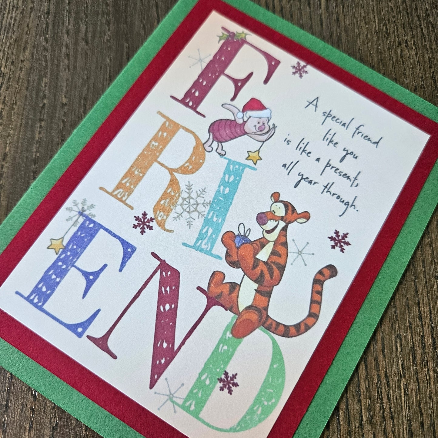A handmade greeting card featuring Winnie the Pooh and Tigger with the word 'FRIEND' spelled out in large decorative letters, placed on a wooden surface.