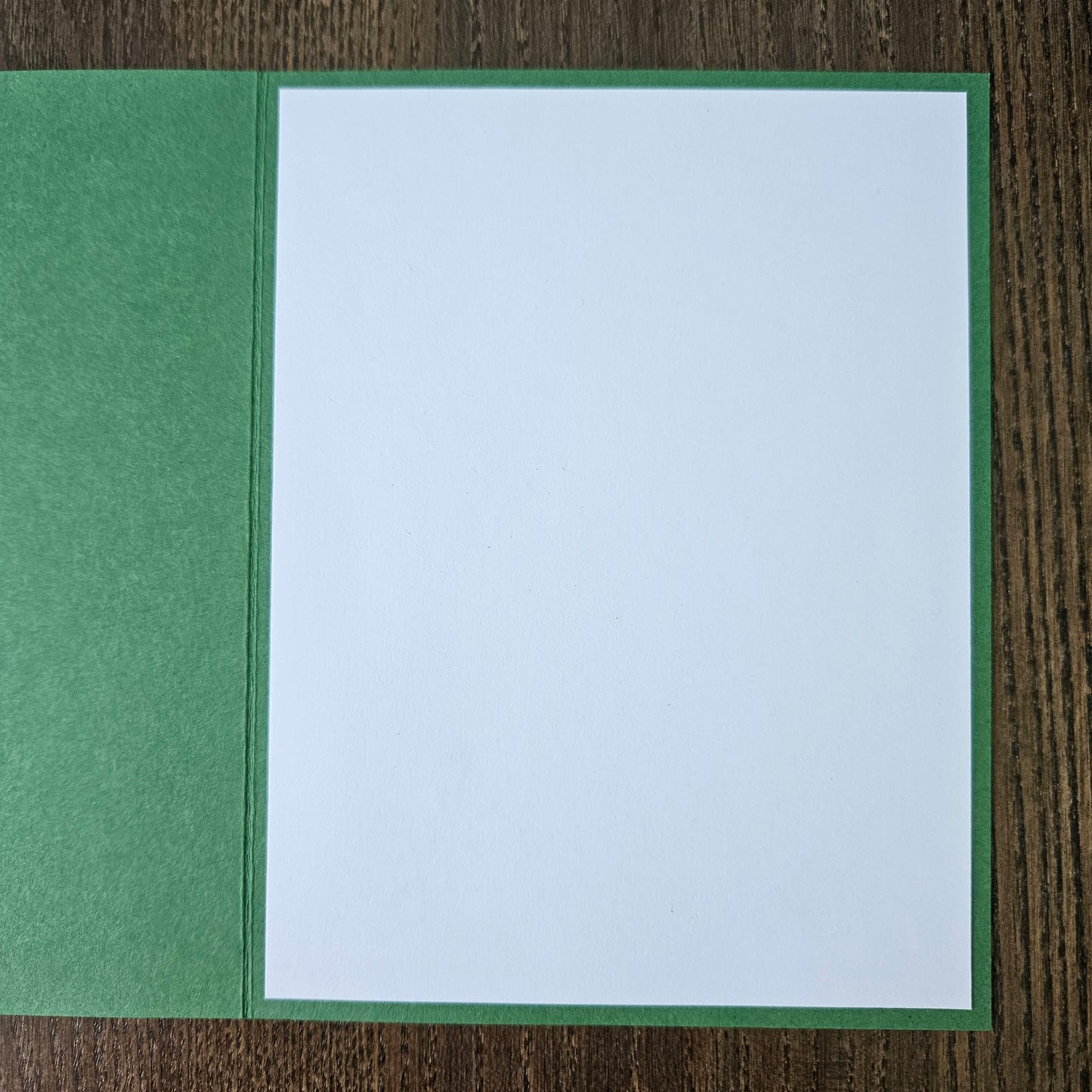A square white card or paper insert with a green border, placed on top of a green textured sheet over a brown wood-grain surface.