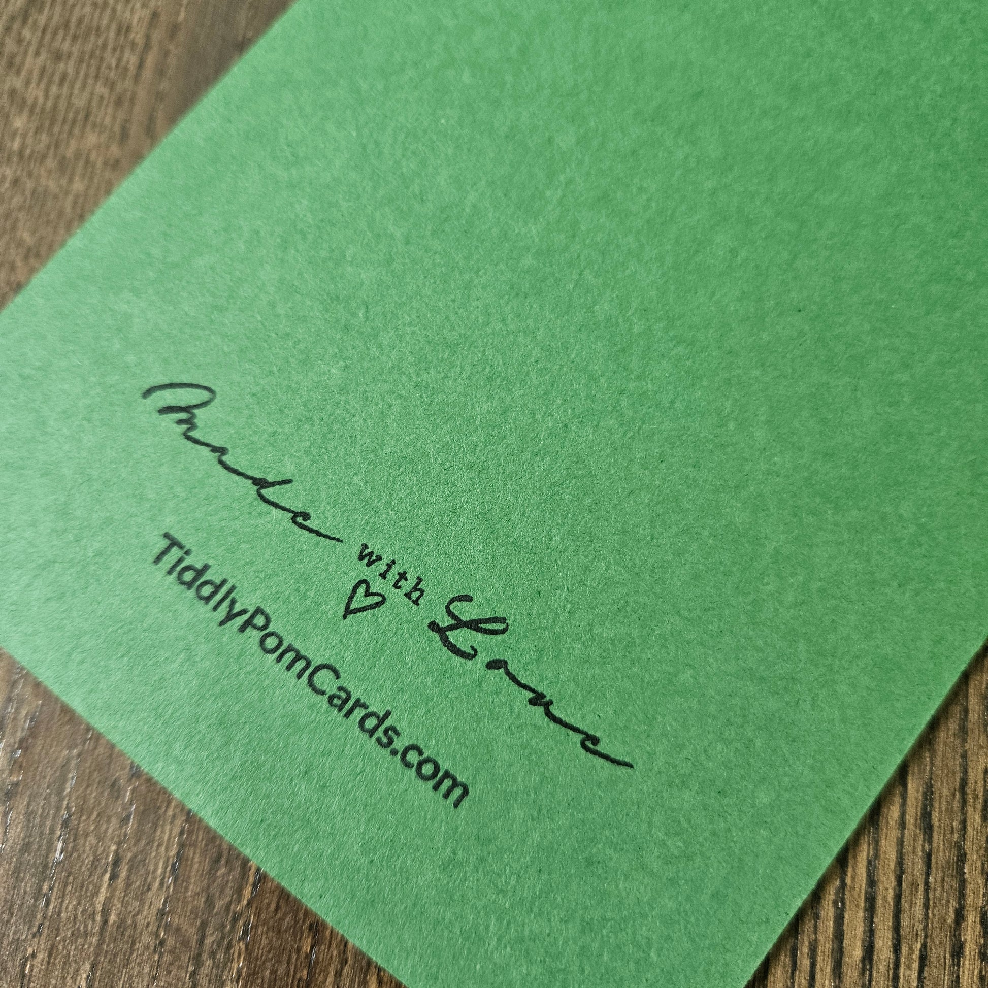 A close-up of a green textured card or paper sheet placed on a wooden surface, printed with the words 'Made with Love' and 'TiddlyPomCards.com' in black script.