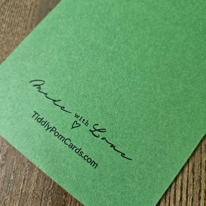 A close-up of a green textured card or paper sheet placed on a wooden surface, printed with the words 'Made with Love' and 'TiddlyPomCards.com' in black script.