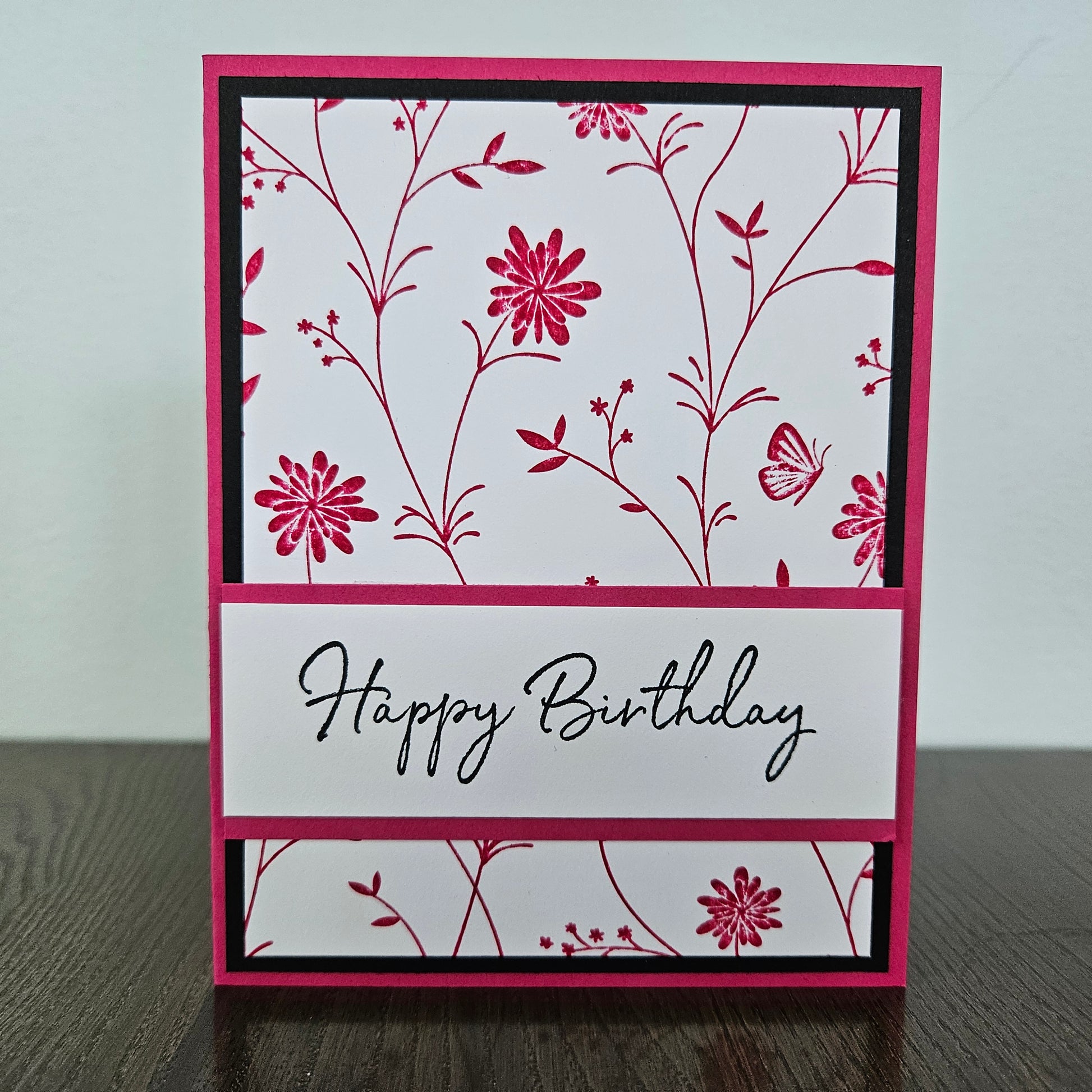 Handmade birthday card for her with pink hand-stamped wildflowers and butterfly design with split banner on black and pink border. Text reads Happy Birthday.