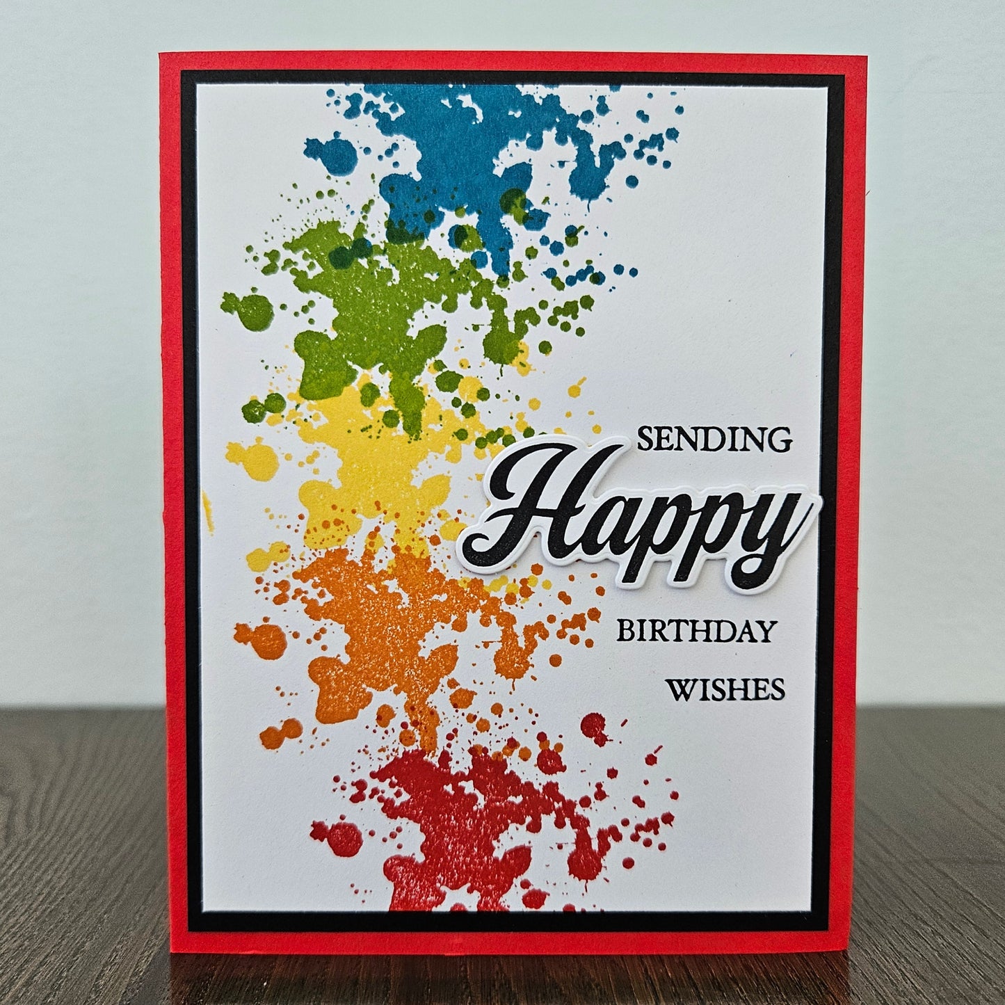 Handmade birthday card with bold rainbow ink splatter design in blue green yellow orange and red with layered die-cut Happy sentiment on black and red border. Text reads Sending Happy Birthday Wishes.