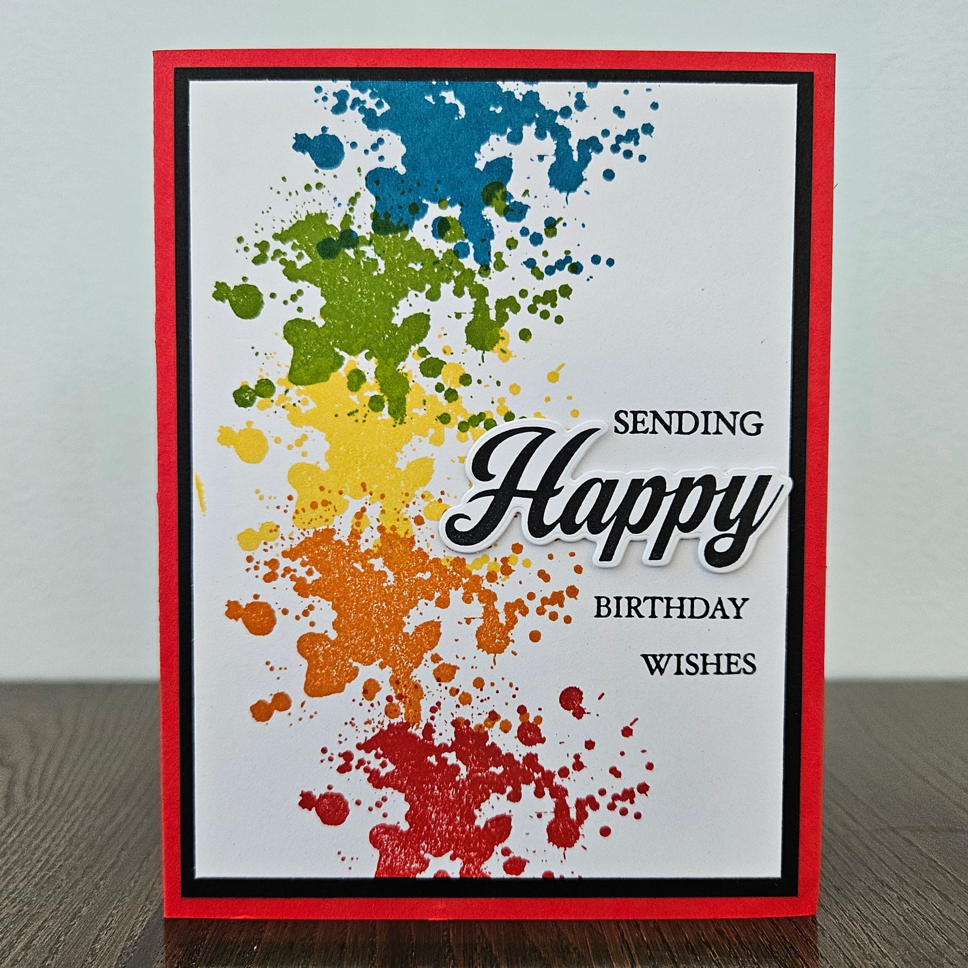 Handmade birthday card with bold rainbow ink splatter design in blue green yellow orange and red with layered die-cut Happy sentiment on black and red border. Text reads Sending Happy Birthday Wishes.