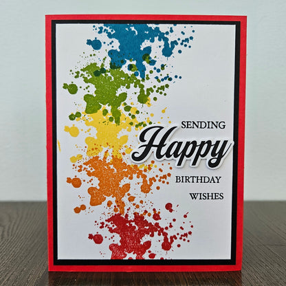 Handmade birthday card with bold rainbow ink splatter design in blue green yellow orange and red with layered die-cut Happy sentiment on black and red border. Text reads Sending Happy Birthday Wishes.