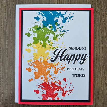 Handmade birthday card with bold rainbow ink splatter design in blue green yellow orange and red with layered die-cut Happy sentiment on black and red border. Text reads Sending Happy Birthday Wishes.
