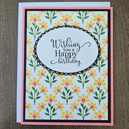 Handmade birthday card for her with hand-stamped yellow daisy field and coral centers with scalloped oval sentiment frame on black and pink border. Text reads Wishing you a Happy Birthday.