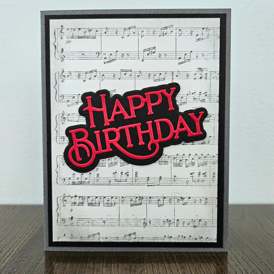 Handmade sheet music birthday card with vintage music score background and bold layered red and black die-cut Happy Birthday sentiment on black and grey border.