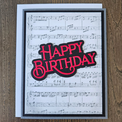 Handmade sheet music birthday card with vintage music score background and bold layered red and black die-cut Happy Birthday sentiment on black and grey border.