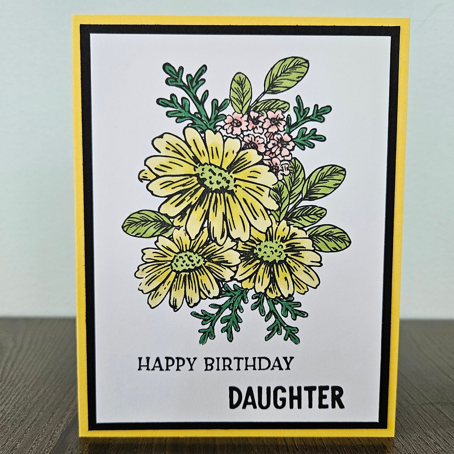 Handmade birthday card for daughter featuring a hand-colored cluster of yellow daisies and tiny pink flowers on a layered white, black, and bright yellow cardstock frame. Bold text reads HAPPY BIRTHDAY DAUGHTER.