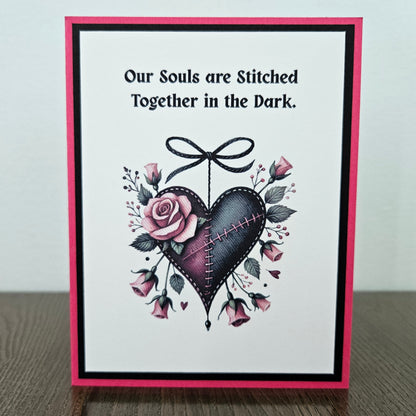 Handmade gothic Valentine card featuring an illustrated purple and black stitched heart with a black bow and pink roses on a hot pink and black layered frame. Text reads Our Souls are Stitched Together in the Dark.