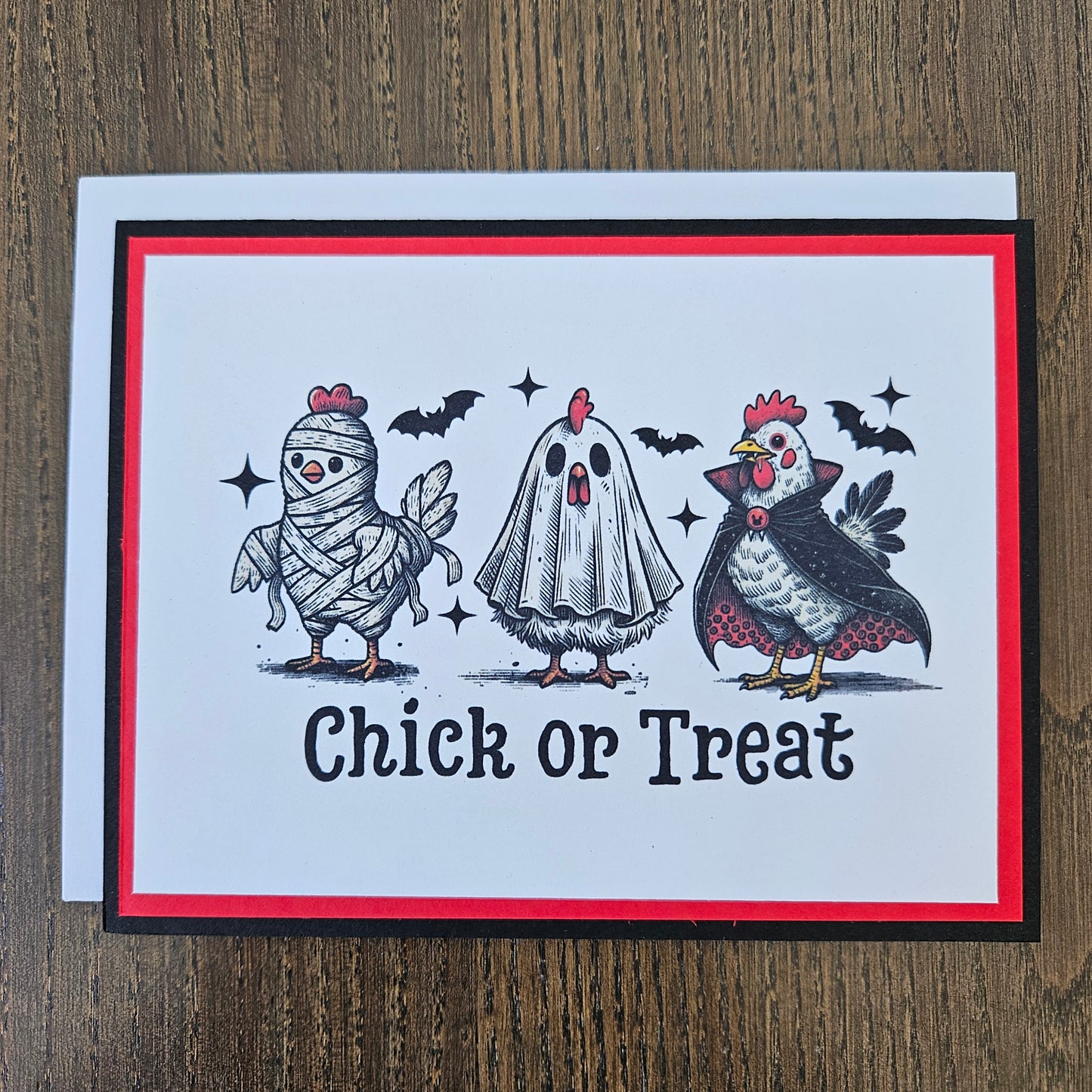 A handmade greeting card featuring a Halloween theme with illustrations of a chicken, a ghost, a mummy, bats, and stars, alongside the text 'Chick or Treat'. The card has a white center, a red border, and a black outer border, resting on a white envelope over a wooden surface.