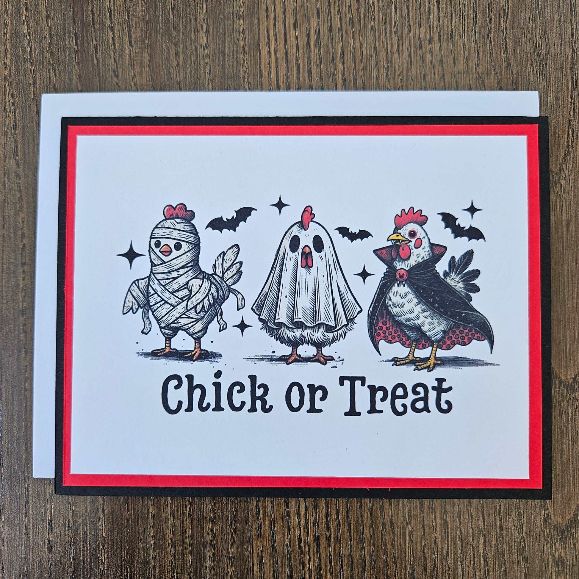 A handmade greeting card featuring a Halloween theme with illustrations of a chicken, a ghost, a mummy, bats, and stars, alongside the text 'Chick or Treat'. The card has a white center, a red border, and a black outer border, resting on a white envelope over a wooden surface.