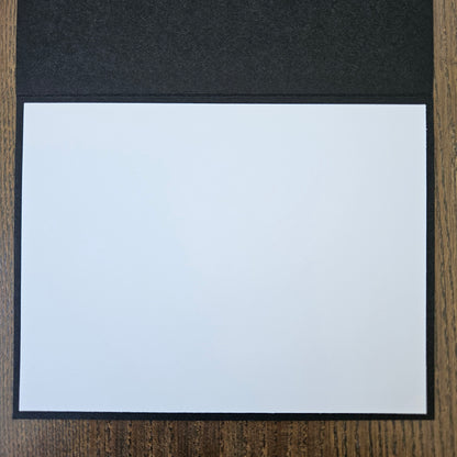 A blank white rectangular card or paper sheet with a black border, placed on a dark textured surface next to a wood-grain background.