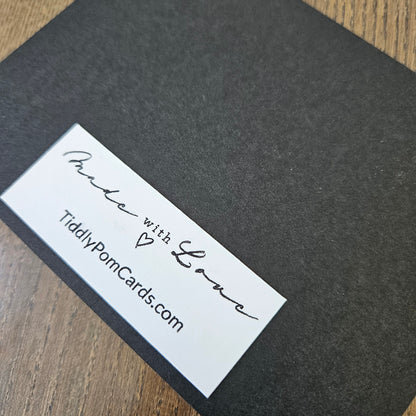 A close-up of a dark charcoal gray textured card or box cover with a white rectangular label that reads 'Made with Love' and 'TiddlyPomCards.com', placed on a wood surface.