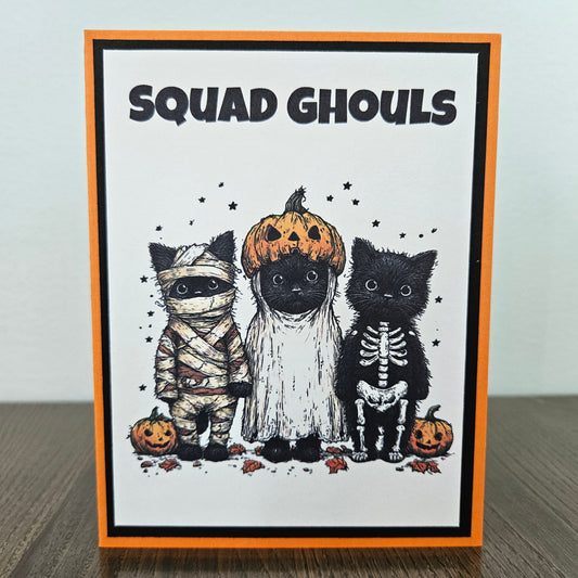 Front of a handmade A2 Halloween card featuring three black cats dressed in mummy, ghost, and skeleton costumes below a bold banner reading Squad Ghouls, layered with cardstock framing.