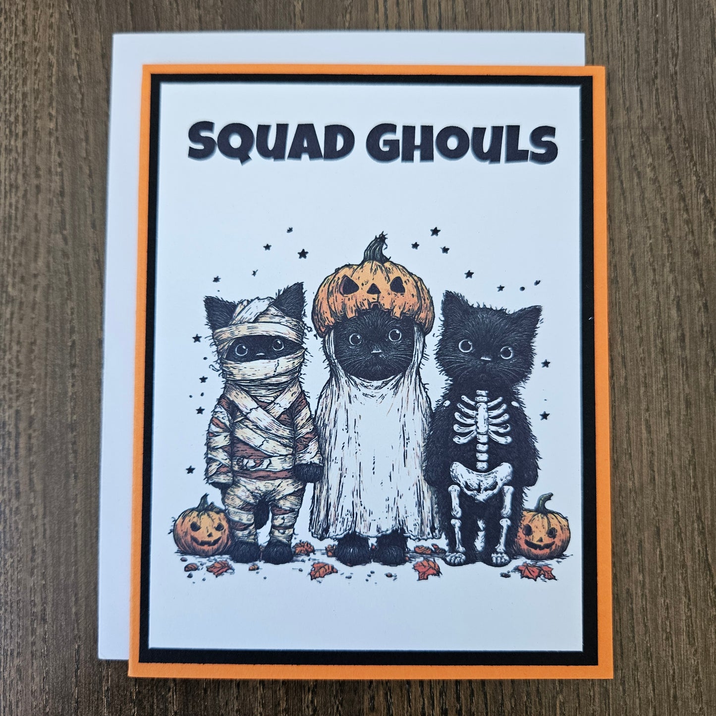 A square greeting card with a Halloween theme, featuring three black cats dressed as pumpkins, skeletons, and a mummy, with the text 'SQUAD GHOULS' on the left. The card has an orange border and is photographed on a brown wooden surface.