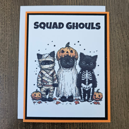 A square greeting card with a Halloween theme, featuring three black cats dressed as pumpkins, skeletons, and a mummy, with the text 'SQUAD GHOULS' on the left. The card has an orange border and is photographed on a brown wooden surface.