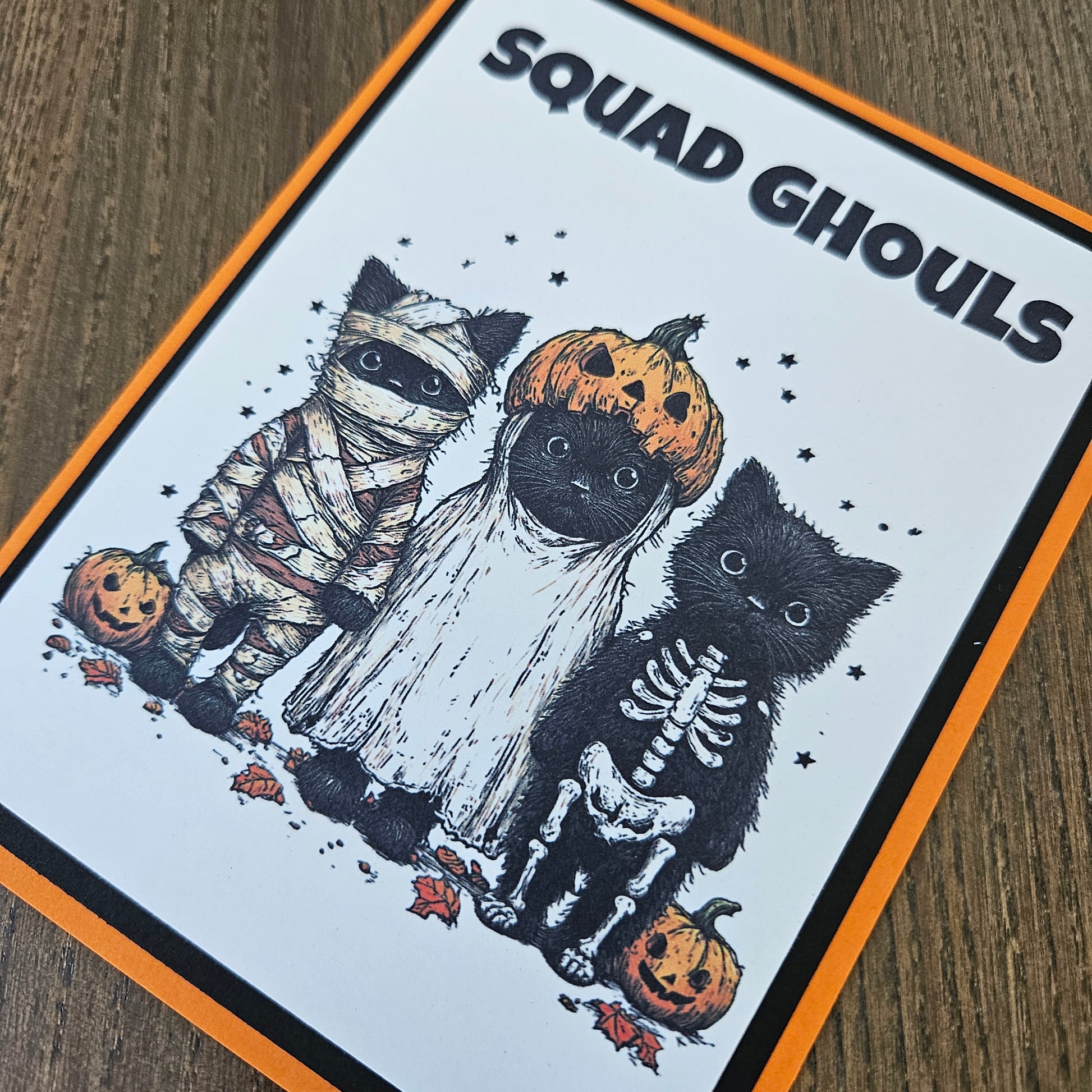 A square printed card or sticker featuring a Halloween illustration with the text 'SQUAD Ghouls' on a white background, bordered in orange and black. The artwork shows black cats dressed as skeletons, a mummy, and pumpkins on a wood surface.