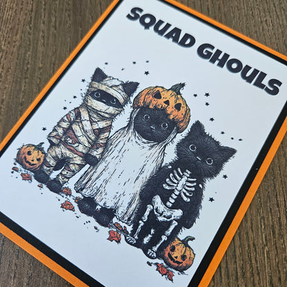 A square printed card or sticker featuring a Halloween illustration with the text 'SQUAD Ghouls' on a white background, bordered in orange and black. The artwork shows black cats dressed as skeletons, a mummy, and pumpkins on a wood surface.