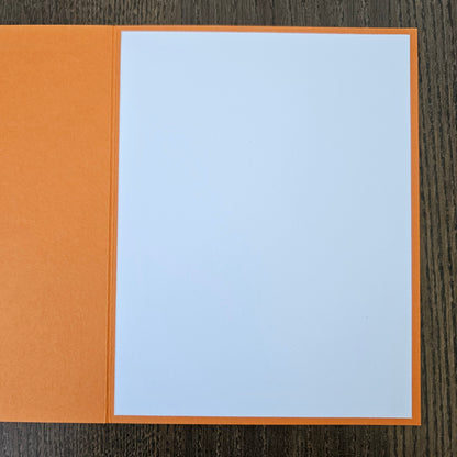A blank white square card or paper insert with a thin orange border, placed on top of an orange paper folder or envelope against a dark wood background.