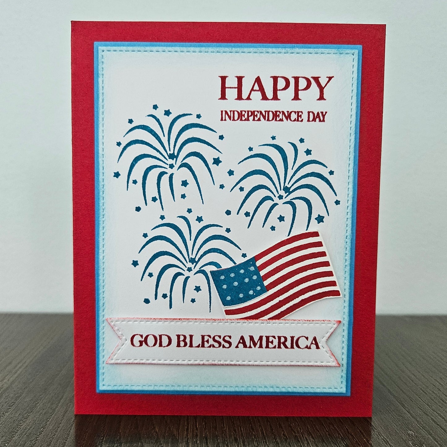 Handmade 4th of July card featuring stamped blue fireworks and a die-cut American flag on a stitched white background panel. The card is framed by a bold red cardstock border with sentiments reading Happy Independence Day and God Bless America.