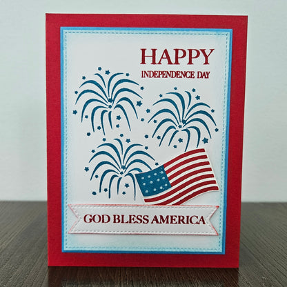 Handmade 4th of July card featuring stamped blue fireworks and a die-cut American flag on a stitched white background panel. The card is framed by a bold red cardstock border with sentiments reading Happy Independence Day and God Bless America.