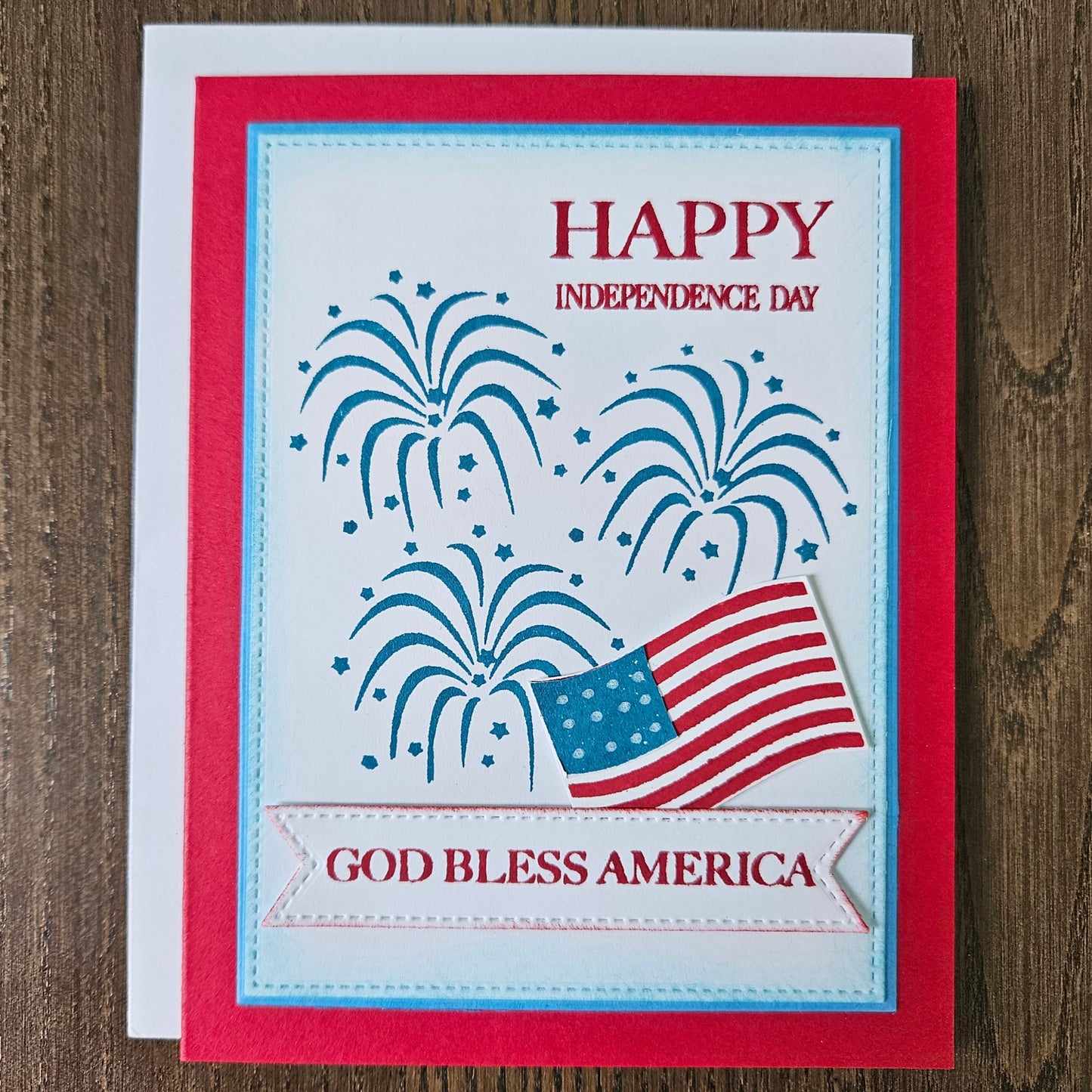 Handmade 4th of July card featuring stamped blue fireworks and a die-cut American flag on a stitched white background panel. The card is framed by a bold red cardstock border with sentiments reading Happy Independence Day and God Bless America.