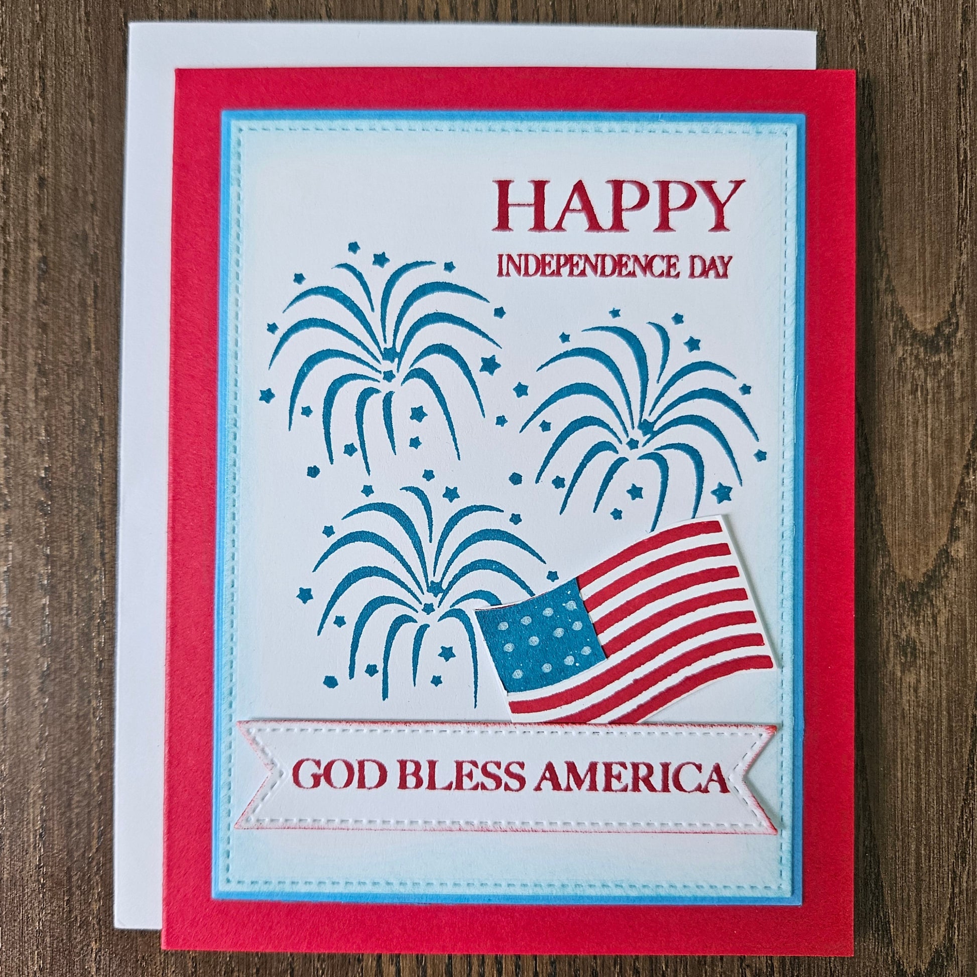 Handmade 4th of July card featuring stamped blue fireworks and a die-cut American flag on a stitched white background panel. The card is framed by a bold red cardstock border with sentiments reading Happy Independence Day and God Bless America.