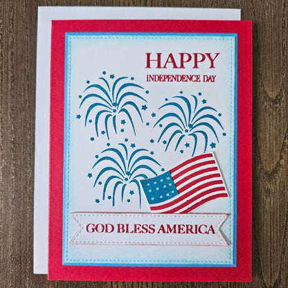 Handmade 4th of July card featuring stamped blue fireworks and a die-cut American flag on a stitched white background panel. The card is framed by a bold red cardstock border with sentiments reading Happy Independence Day and God Bless America.