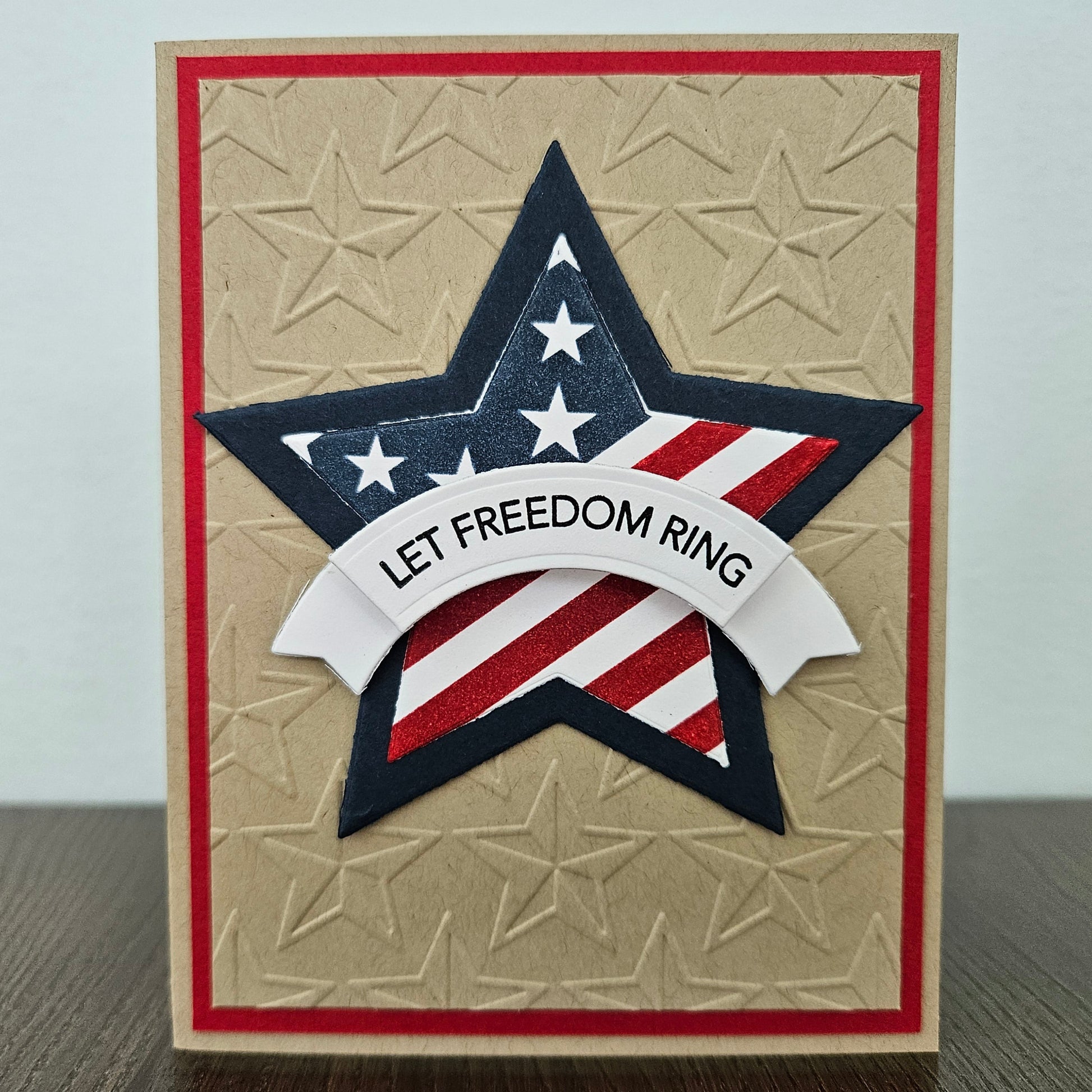 Handmade 4th of July card featuring a large layered star with an American flag design and a white dimensional banner reading Let Freedom Ring. The star is centered on a rustic kraft paper background deeply embossed with a repeating star pattern and framed by a thin red border layer.