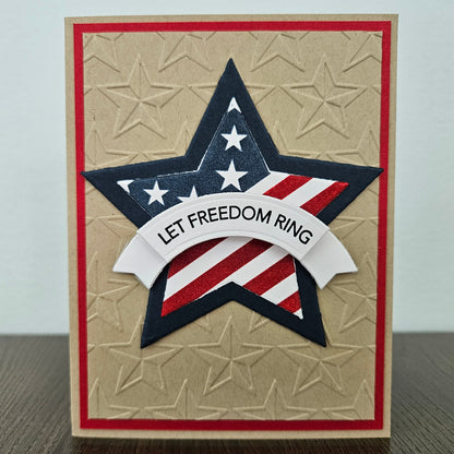 Handmade 4th of July card featuring a large layered star with an American flag design and a white dimensional banner reading Let Freedom Ring. The star is centered on a rustic kraft paper background deeply embossed with a repeating star pattern and framed by a thin red border layer.